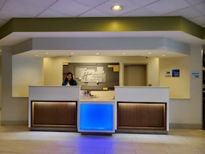 Un mostrador de recepción con una pantalla digital, un cartel de Holiday Inn Express & Suites y un letrero de Marketplace que indica acceso las 24 horas.
