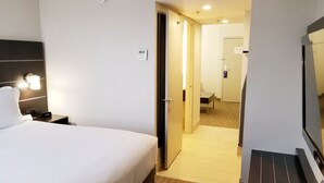 A hotel room with a bed, bedside table, and a nightstand with a clock. There is a door leading to a bathroom and a hallway.