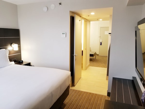 Ein Hotelzimmer mit einem Bett, einem Nachttisch und einem Beistelltischchen mit einer Uhr. Eine Tür führt zu einem Badezimmer und einem Flur.