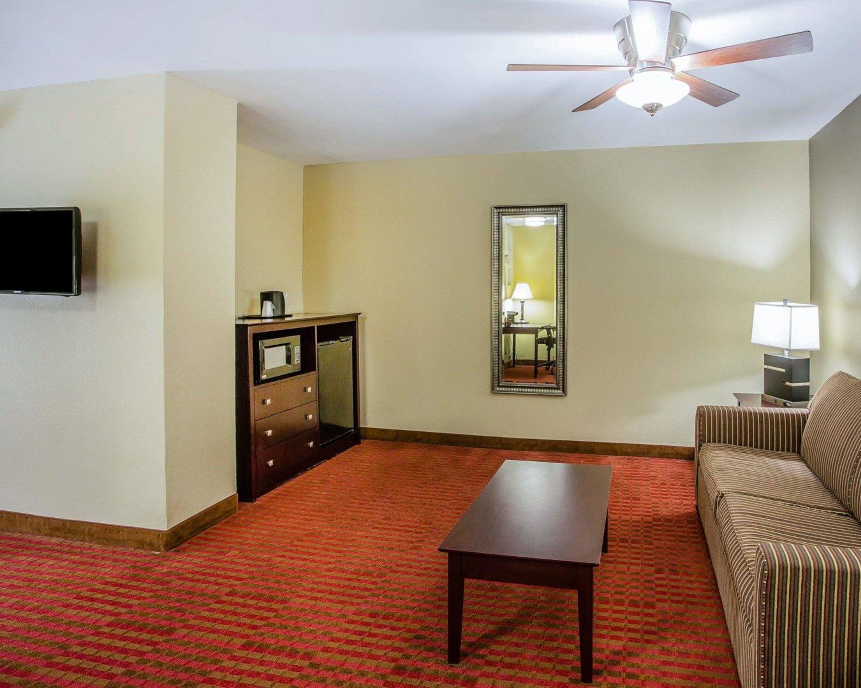 Une chambre d’hôtel équipée d’un téléviseur à écran plat, d’un bureau, d’un canapé, d’un miroir, d’un ventilateur de plafond et d’un sol recouvert d’une moquette rouge.