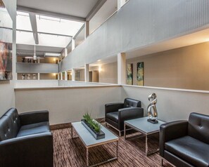 Um hall de hotel moderno com teto alto, área de estar central com poltronas e mesas de couro, e obras de arte nas paredes.