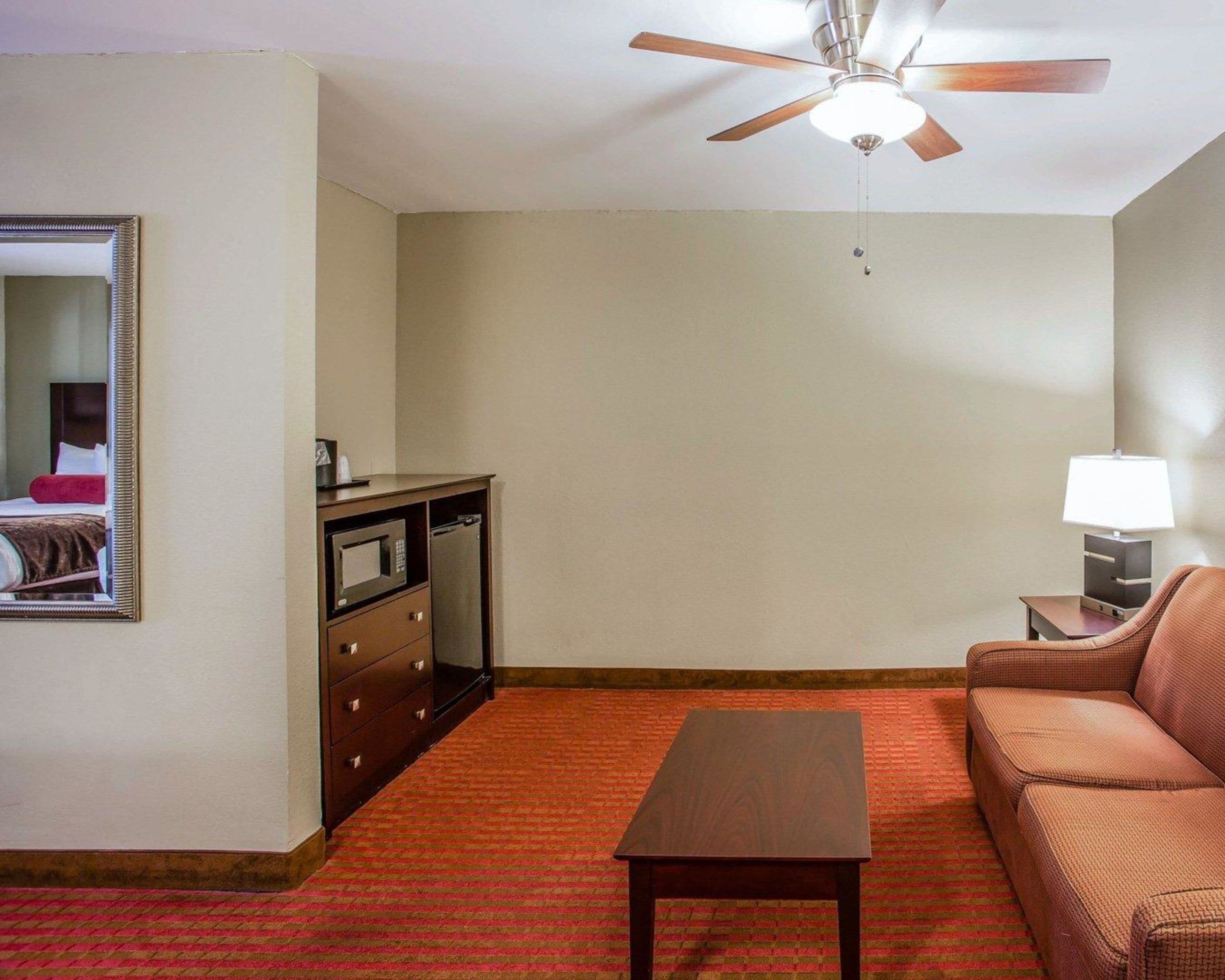 Habitación de hotel con cama, cómoda de madera, microondas, cafetera, lámpara, alfombra roja y ventilador de techo.