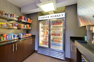 A corner market with shelves stocked with snacks and beverages, a refrigerated section, and a stainless steel range hood.