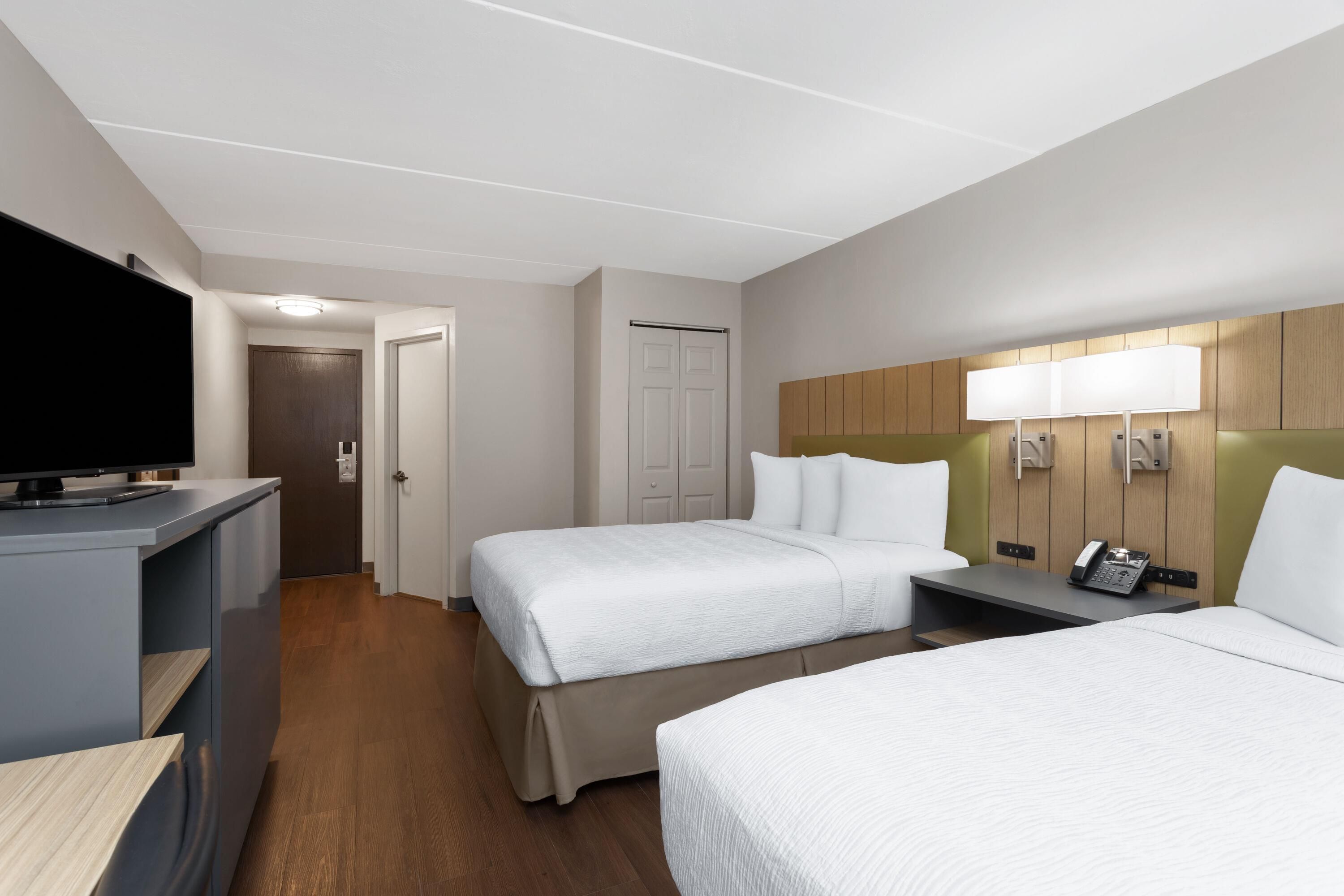 A hotel room with two beds, a desk with a flat-screen TV, a bedside table with a phone, and a wall-mounted lamp.