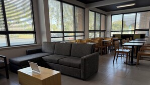 Una habitación moderna con un sofá seccional gris, mesas y sillas de madera, ventanas grandes con persianas y suelo de baldosas.