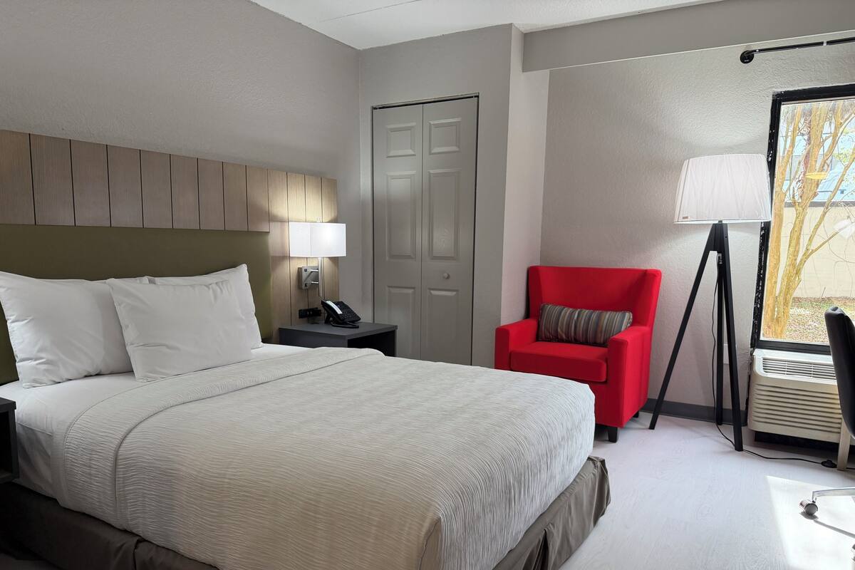 A hotel room with a large bed, a desk with a phone, a red chair, a standing lamp, and a window with a view.