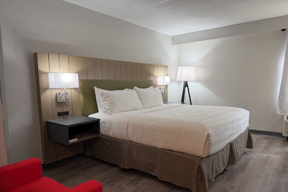 A hotel room with a bed, bedside lamps, a nightstand, a red chair, and a wooden floor.