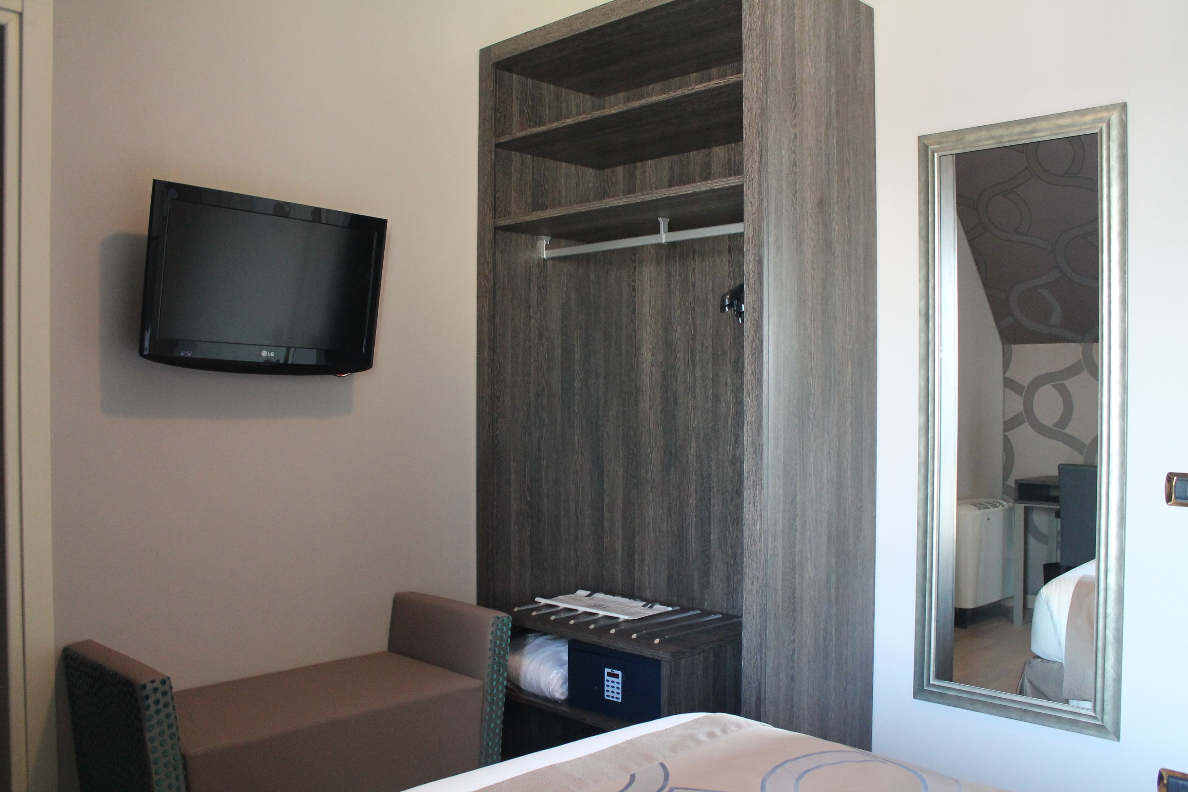 A hotel room with a flat-screen TV mounted on the wall, a bed with a headboard, a seating area with a chair, a wardrobe with a mirror, and a small table with a lamp.