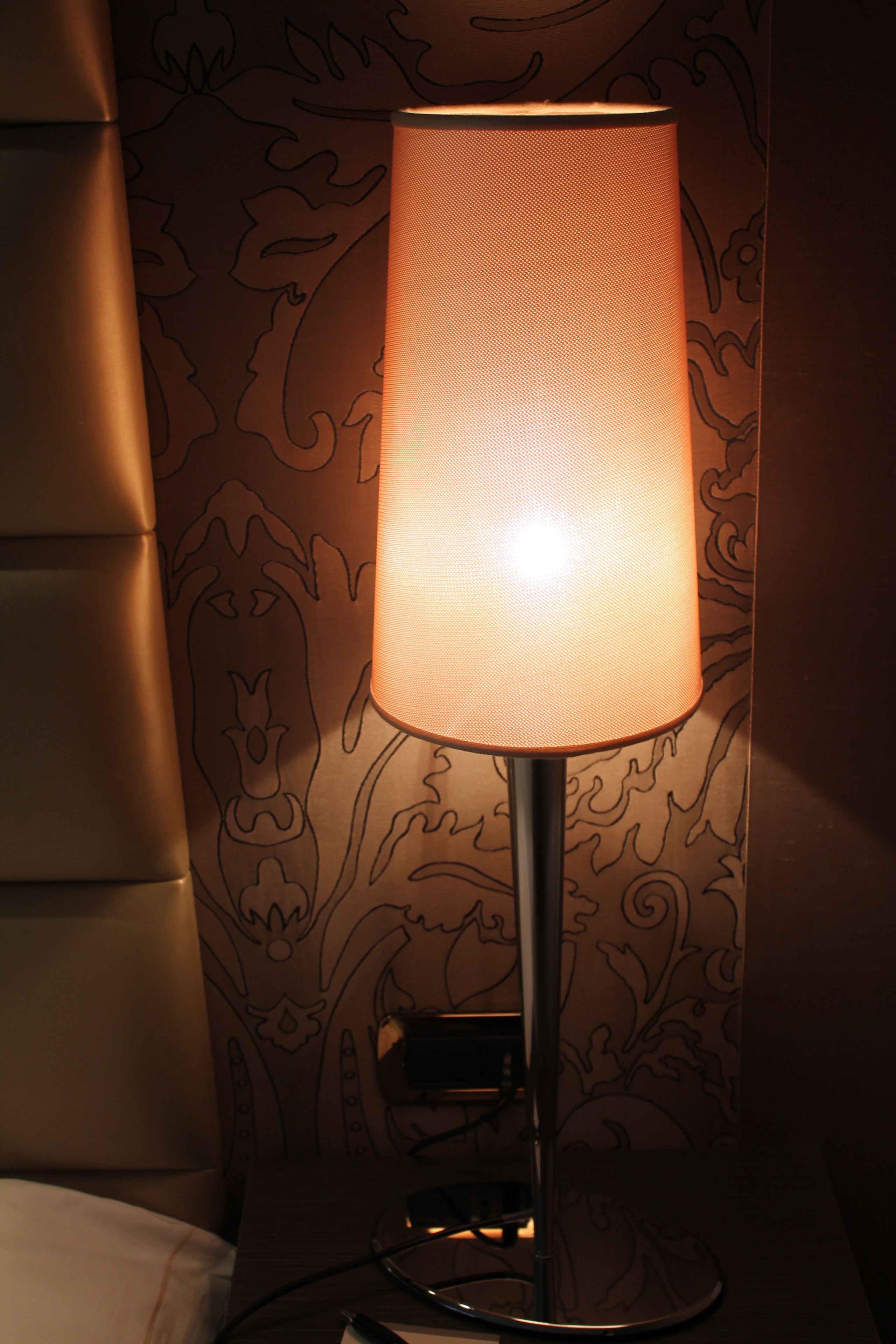 A bedside lamp with a textured shade, a power outlet, and a wall with a floral pattern.