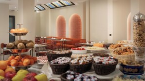 Un bufé con diversas frutas, pasteles y pan. Hay cesteras de mimbre, un bol de frutas y un recipiente grande de granola. La habitación tiene techos altos con tragaluces y asientos color naranja al fondo.