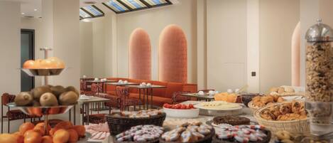 A buffet with various fruits, pastries, and bread. There are wicker baskets, a fruit bowl, and a large container of granola. The room has a high ceiling with skylights and orange seating in the background.