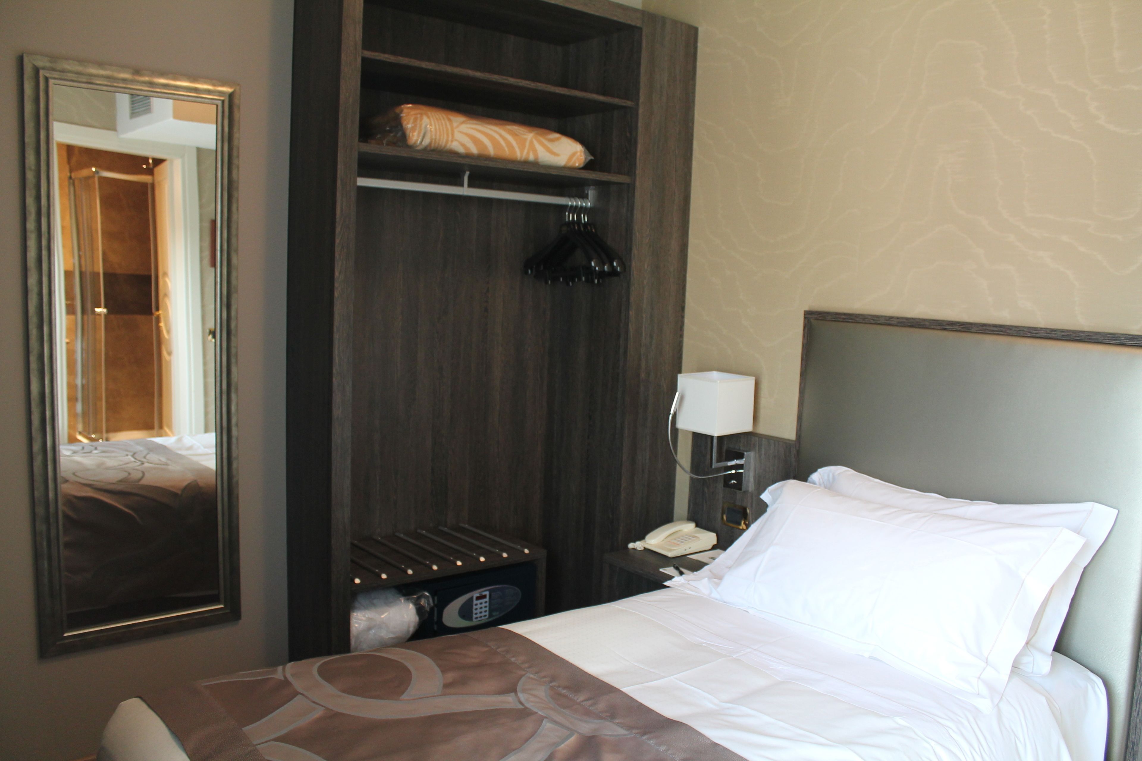 Une chambre d’hôtel équipée d’un lit, d’une armoire, d’un miroir et d’une table de chevet avec une lampe et un téléphone.