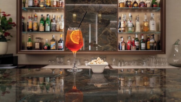 Zona de bar con una copa de cóctel rojo, un cuenco de frutos secos y estantes llenos de botellas de licor.