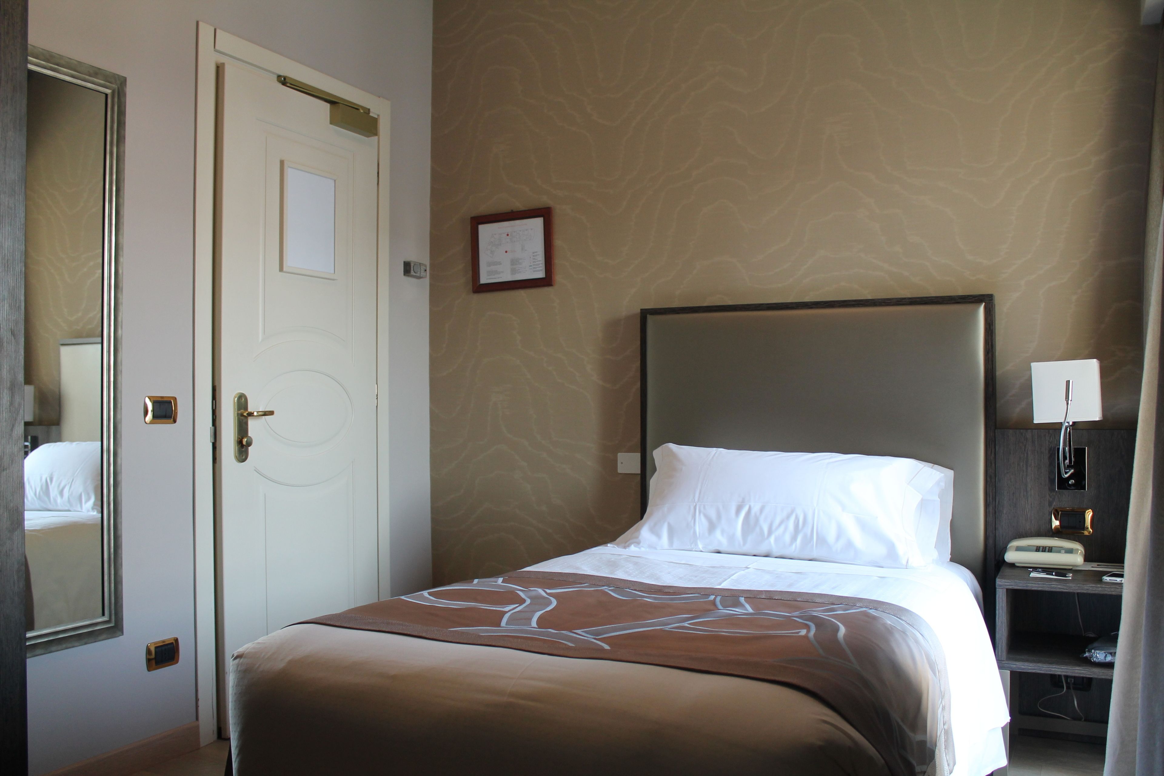 Een hotelkamer met een bed, nachtkastje, spiegel, deur en een muur met een patroon.