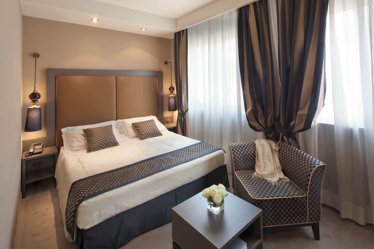 Een hotelkamer met een groot bed, een stoel, een tafeltje met een bloemenvaas en gordijnen.
