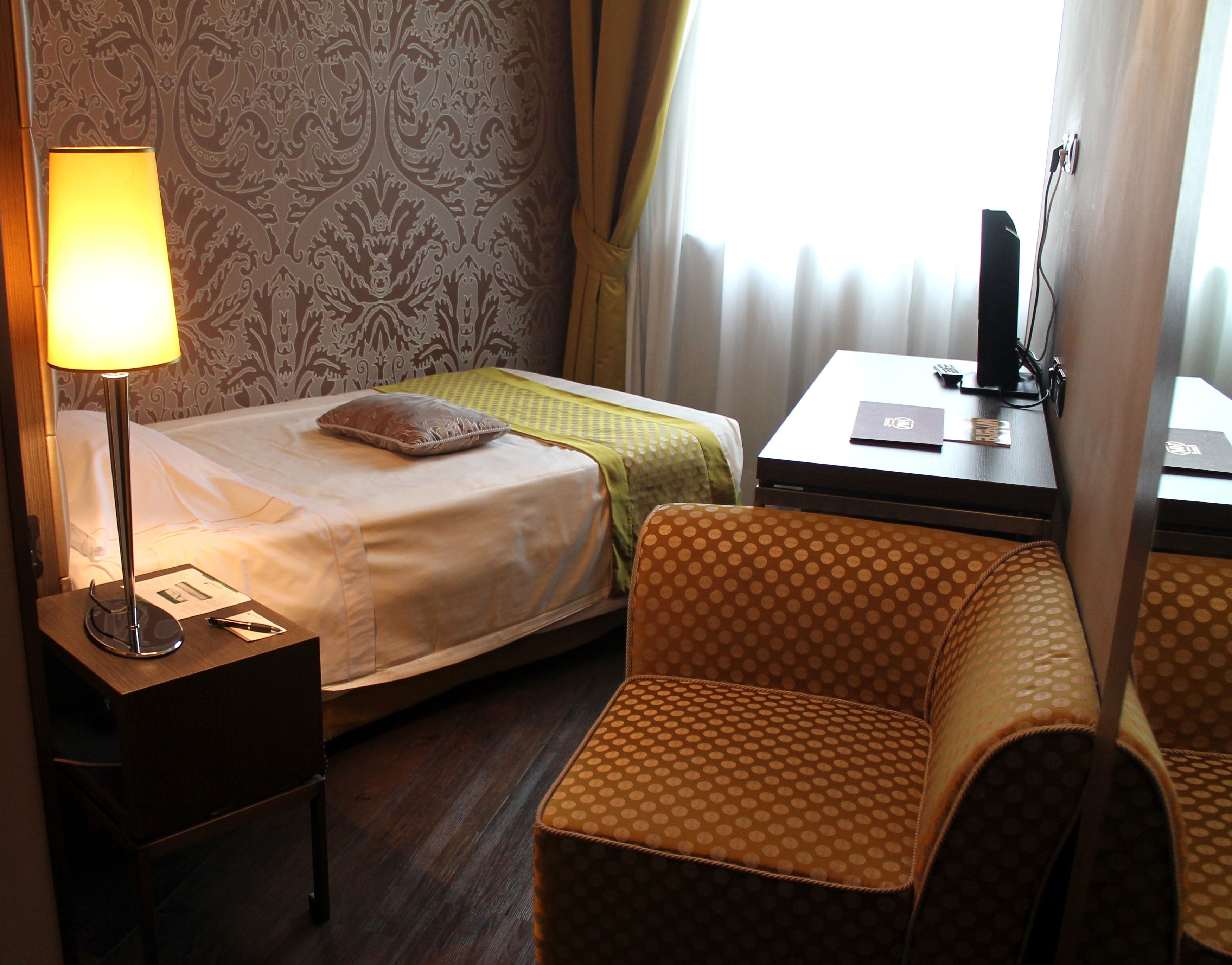 A hotel room with two beds, a desk with a lamp, a chair, and a view of the outside through a window.
