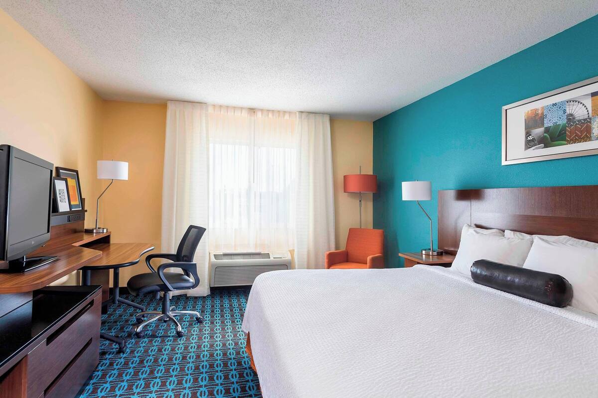 Ein Hotelzimmer mit einem Bett, einem Schreibtisch mit Computer, einem Sessel und einer Klimaanlage sowie einem Fernseher.