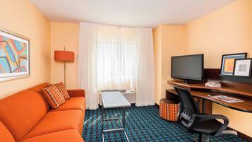 Quarto de hotel com sofá, escrivaninha com computador, televisão e carpete estampado.