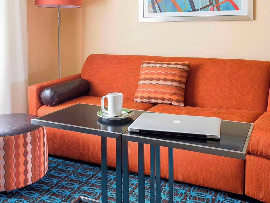 Una habitación de hotel moderna con un sofá naranja, una mesa de centro de vidrio con un ordenador portátil, una alfombra estampada y una lámpara de pie.