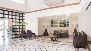 A spacious lobby with a high ceiling, a seating area with purple chairs, a sofa, and a coffee table. There is a large window with a view of the outside, and a balcony with potted plants. - iStay Hotel Ciudad Juarez (Ciudad Juárez)