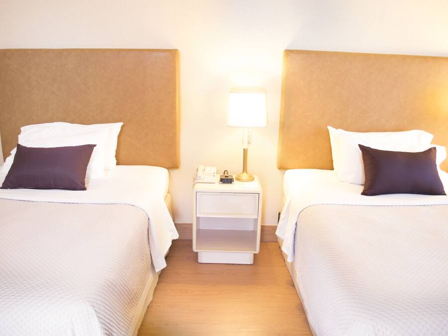 Quarto de hotel com duas camas, cada uma com cabeceira, mesa de cabeceira com abajur e gavetinha.