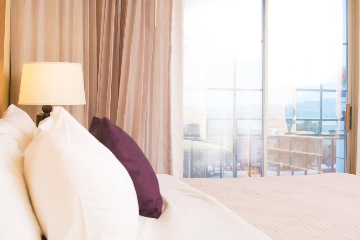 A hotel room with a large bed, a nightstand with a lamp, and a view of a balcony with a cityscape.