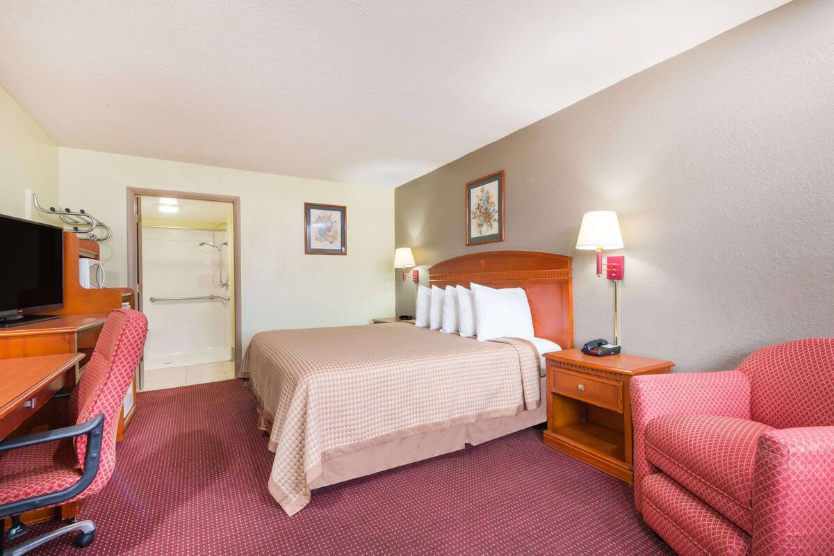A hotel room with a bed, a desk with a computer, a chair, a TV, and a lamp.