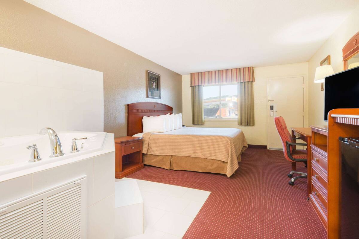 A hotel room with a bed, a desk, a chair, a TV, and a bathroom with a sink and mirror.