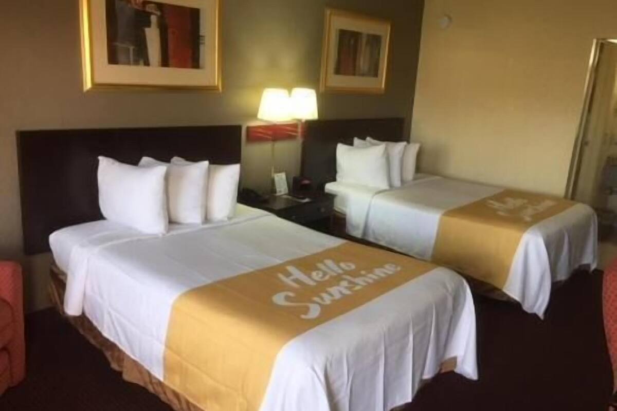 A hotel room with two beds, each with a 'Hello Sunshine' bedspread, a nightstand with a lamp, and framed pictures on the wall.