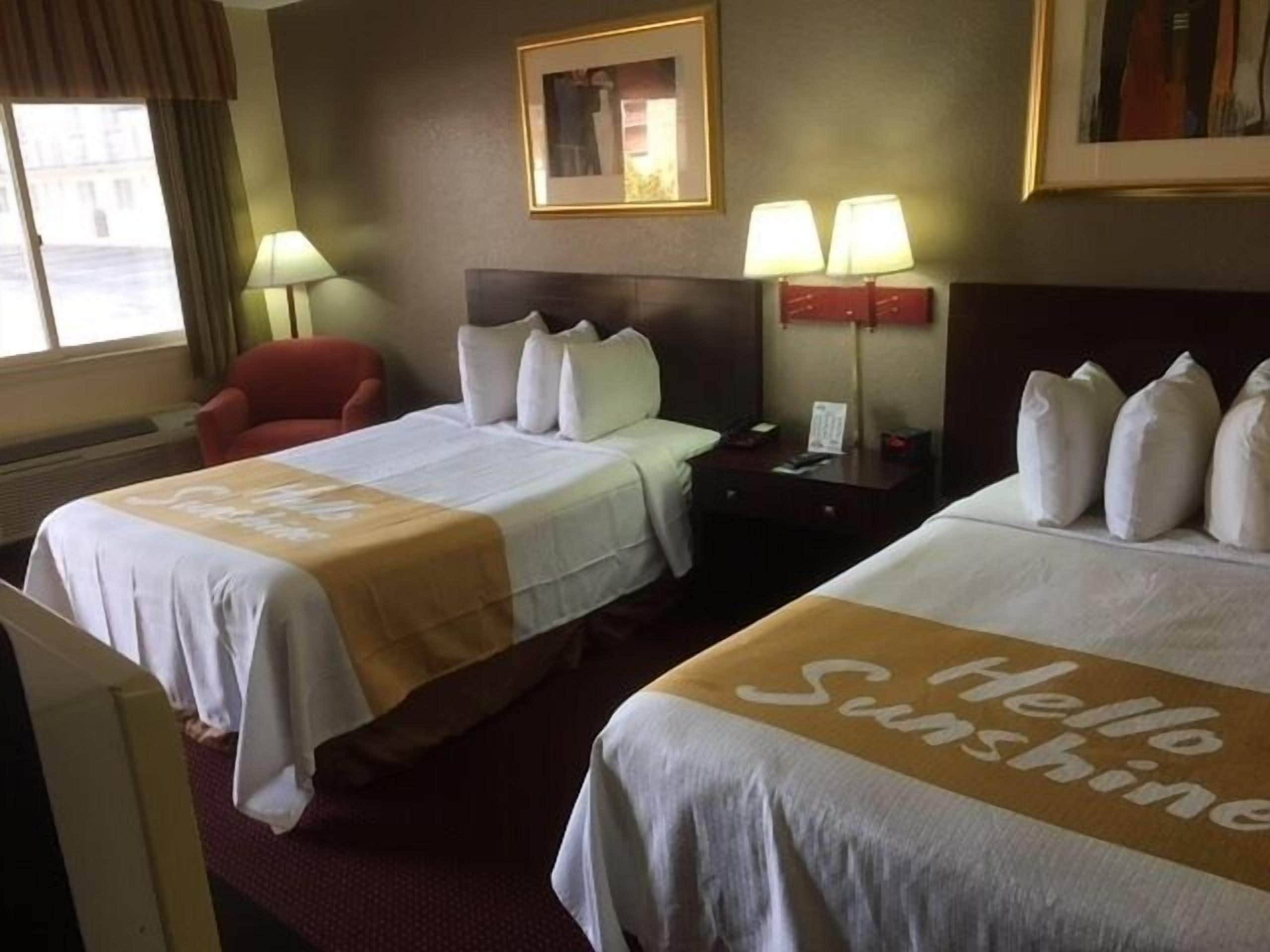 A hotel room with two beds, a red chair, a nightstand with lamps, and a framed picture on the wall.