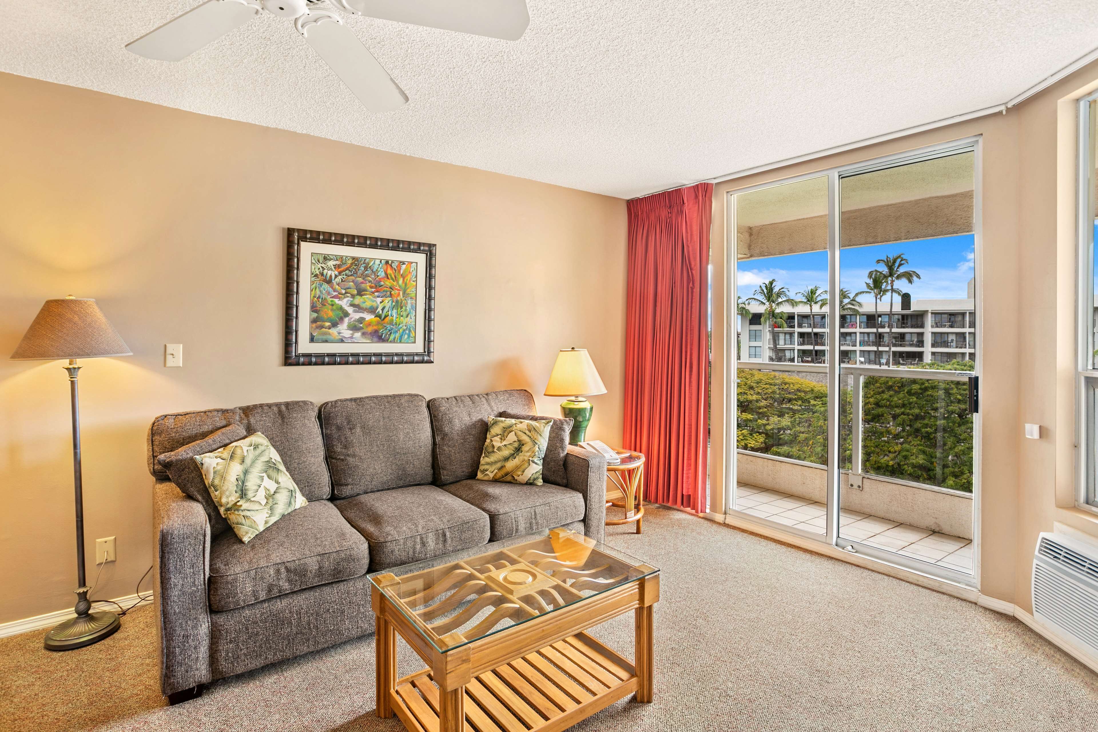 A living room with a gray sofa, a glass coffee table, a floor lamp, a painting, and a view of a building with palm trees through large sliding glass doors.