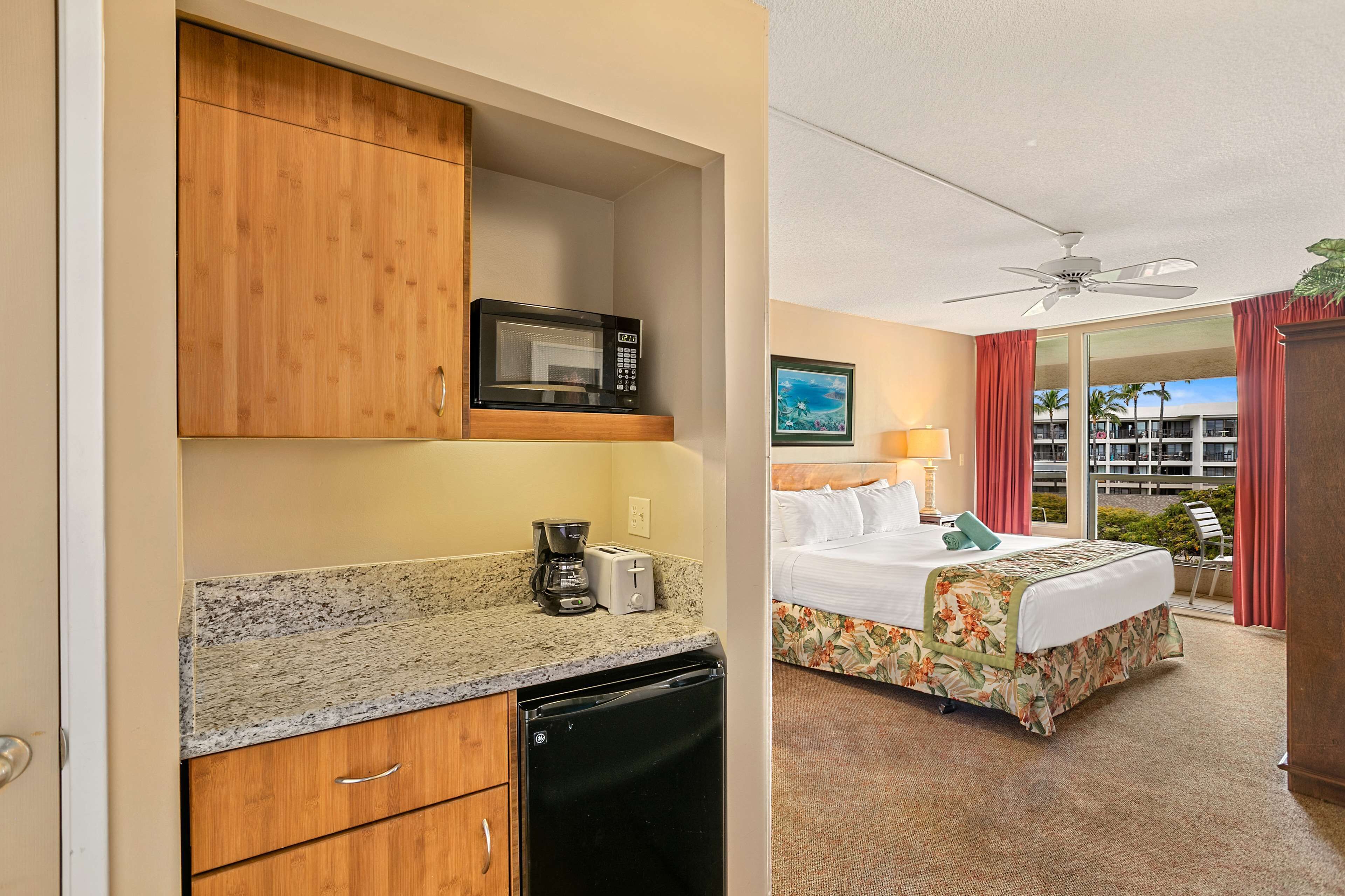 A hotel room with a kitchenette, a bed with floral bedding, a ceiling fan, and a view of palm trees.