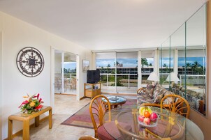A spacious living area with a large window, a television, a dining table with fruit, and a sofa.