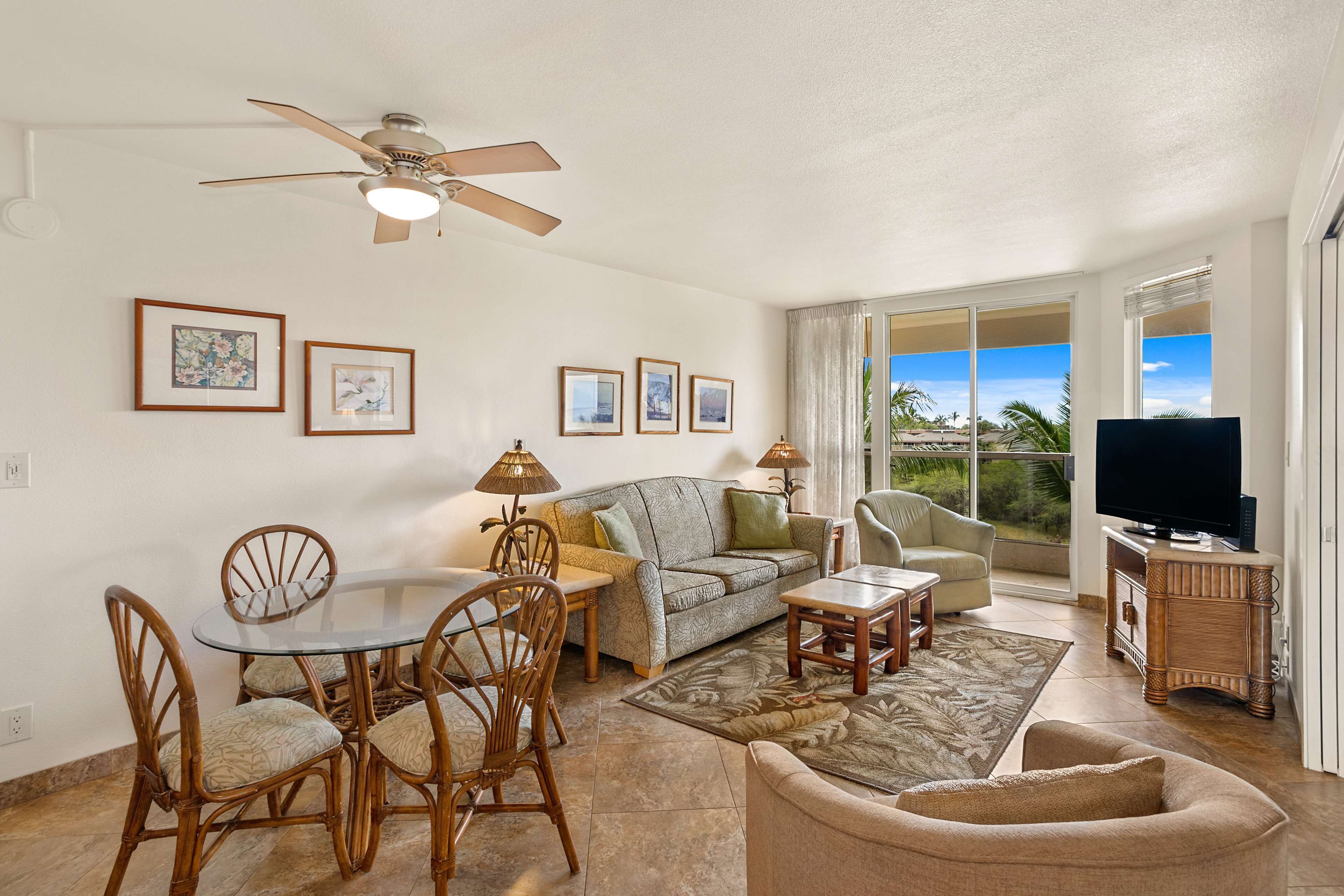 A spacious living room with a ceiling fan, a dining table with chairs, a sofa, a TV stand, and large windows offering a view of palm trees.