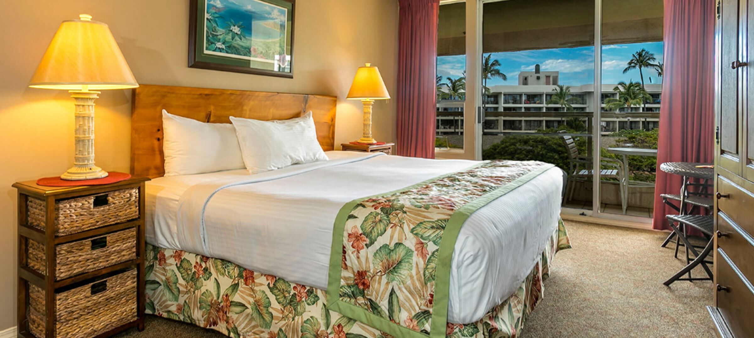 Habitación de hotel con cama, dos lámparas de noche, una mesita de noche de madera con cestas, y un balcón con vistas a palmeras y edificios.
