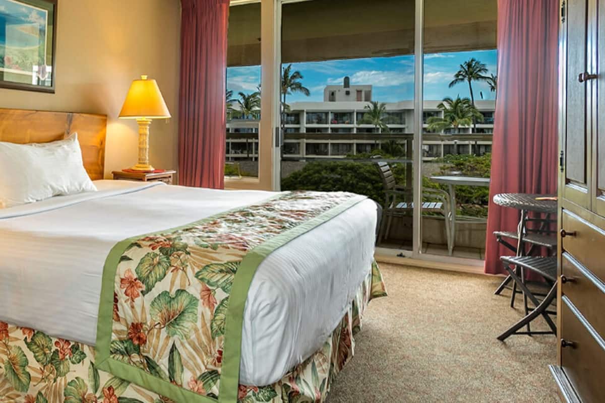 Habitación de hotel con cama, dos lámparas de noche, una mesita de noche de madera con cestas, y un balcón con vistas a palmeras y edificios.