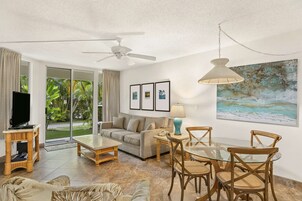 A spacious living room with a sofa, coffee table, and dining table with chairs. There is a ceiling fan, a TV on a stand, and a painting on the wall.