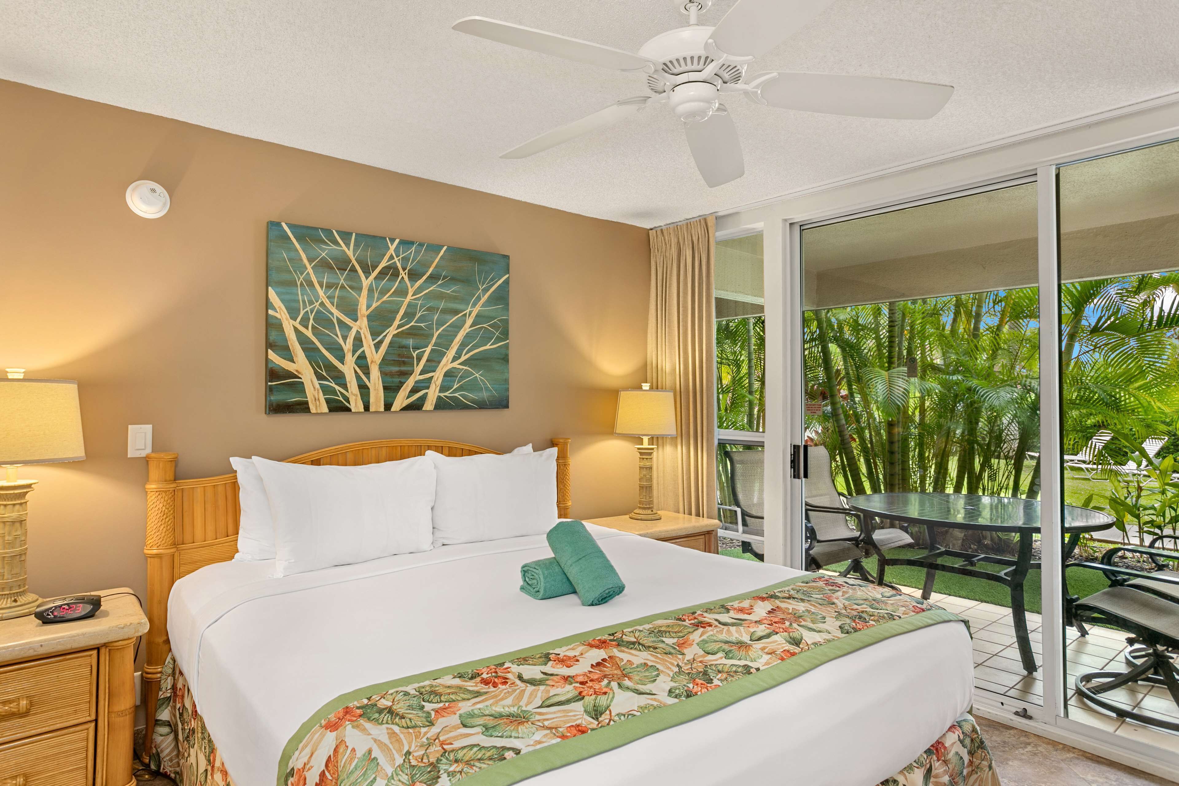A hotel room with a bed, a ceiling fan, a painting of trees, a balcony with a glass door, and a view of greenery.