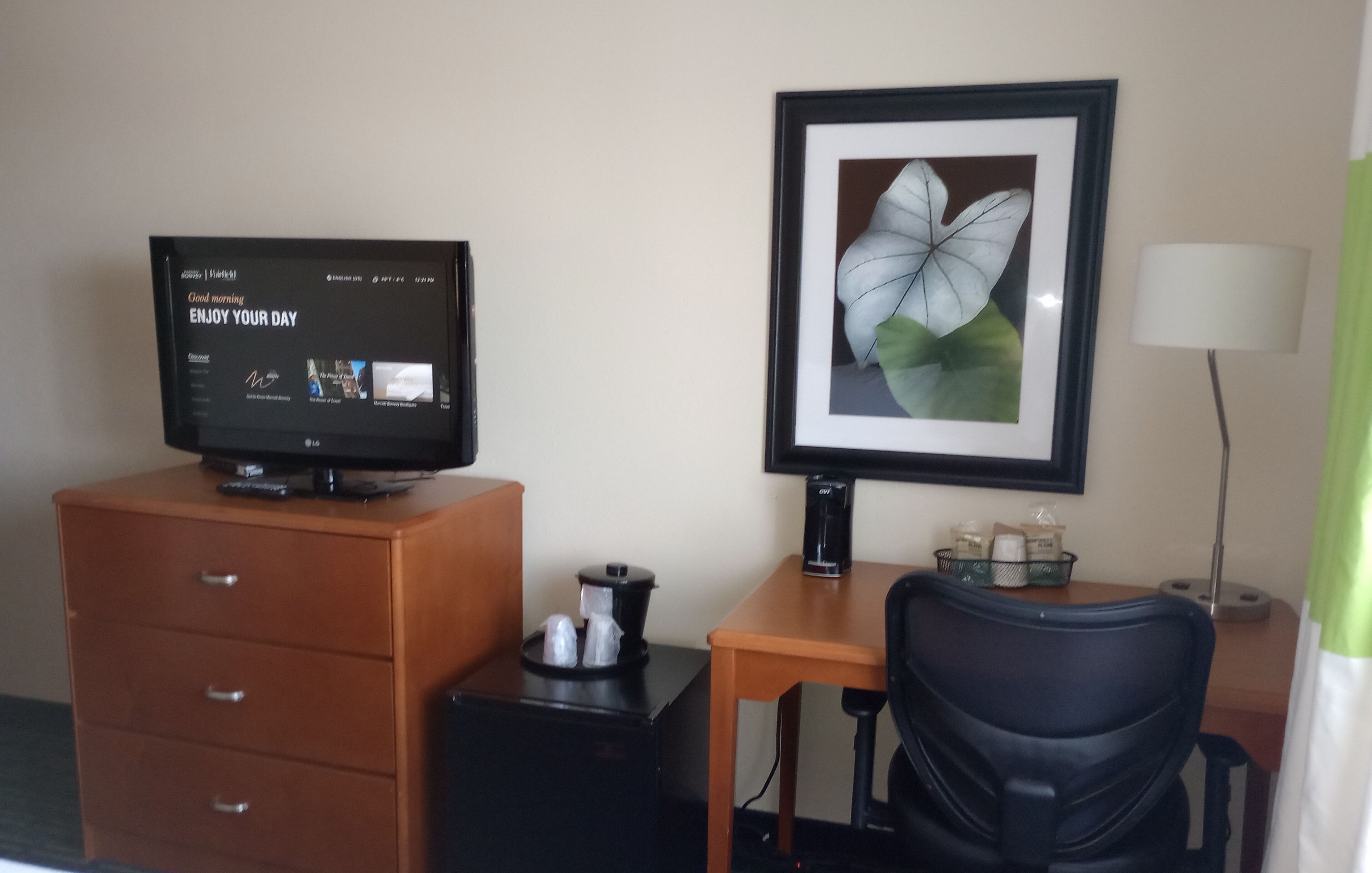 A hotel room with a television on a wooden dresser, a framed picture on the wall, a desk with a lamp, and a chair.