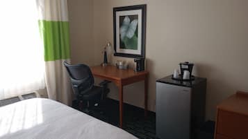 Habitación de hotel con cama, escritorio, silla, cafetera, refrigerador y un cuadro en la pared.