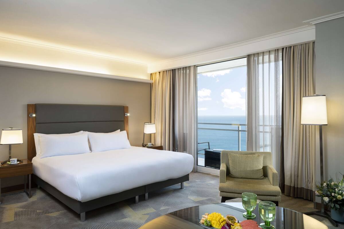 A hotel room with a large bed, a sofa, a balcony with a view of the sea, and a table with a vase of flowers.