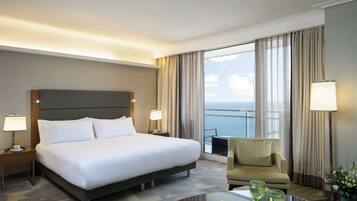 Habitación de hotel con una cama grande, un sofá, un balcón con vistas al mar y una mesa con un jarrón de flores.