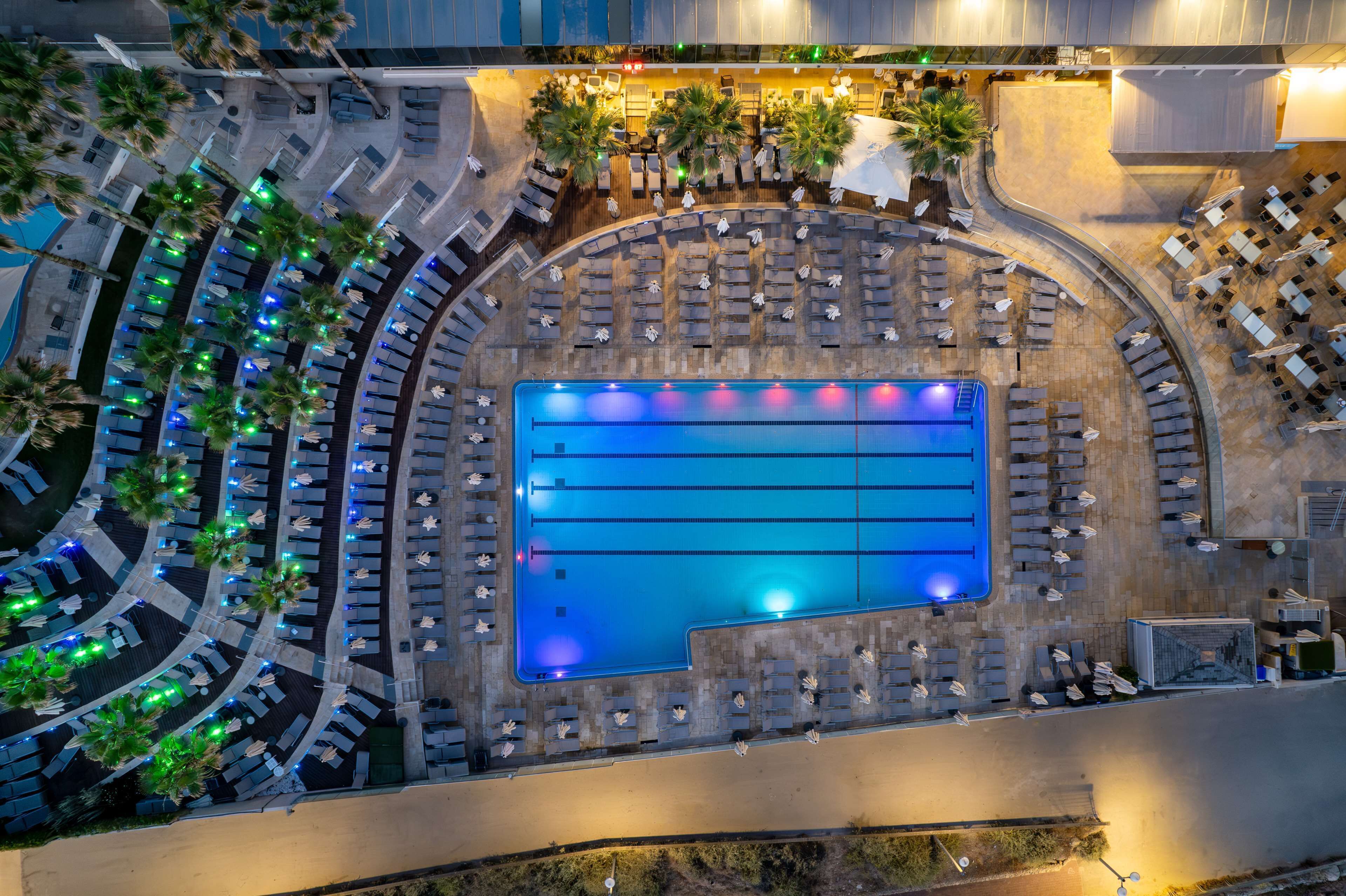 An aerial view of a hotel with a large swimming pool, multiple lounge areas, and palm trees.