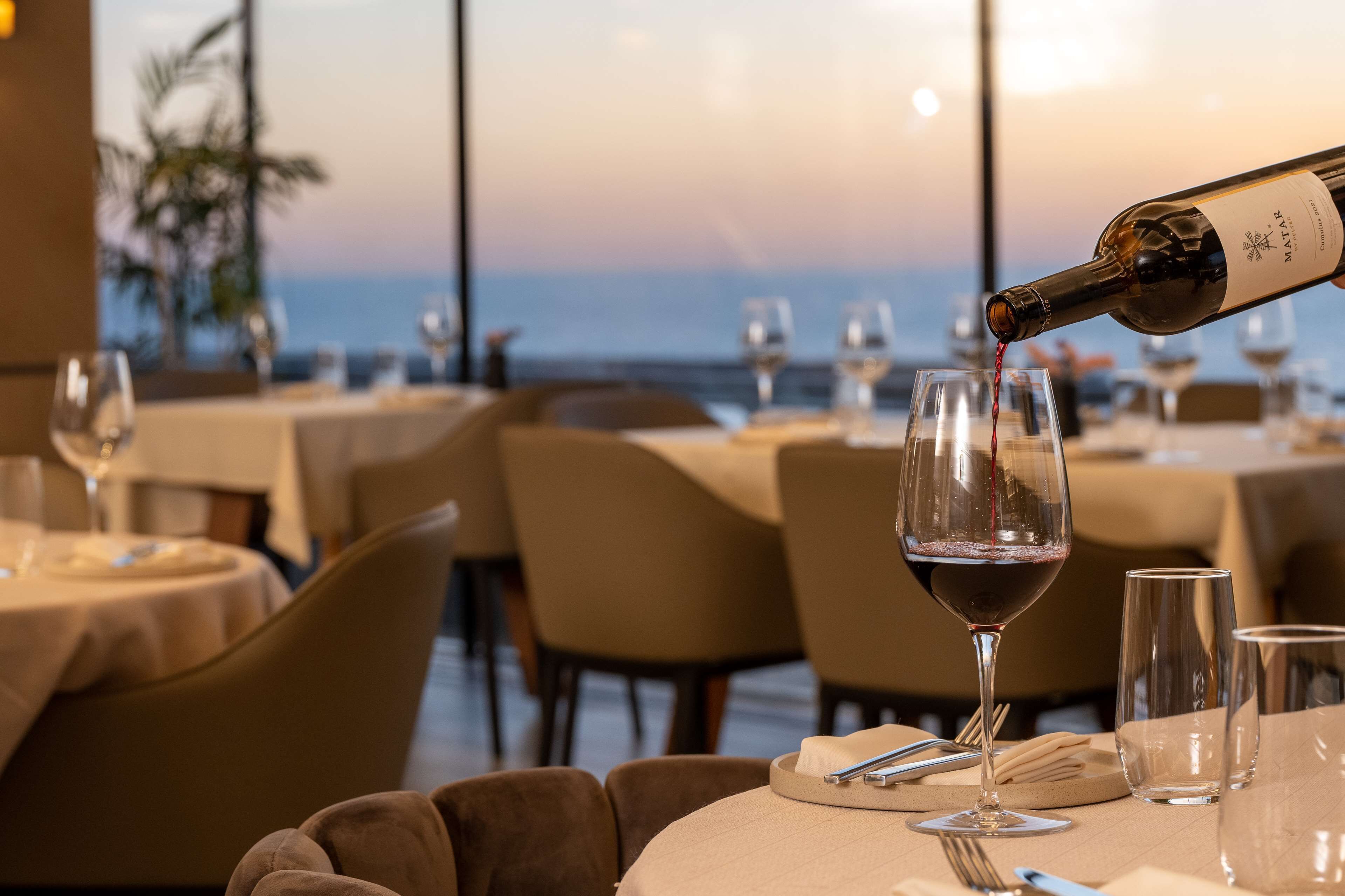 A restaurant with tables set for dining, a bottle of wine being poured into a glass, and a view of the ocean.