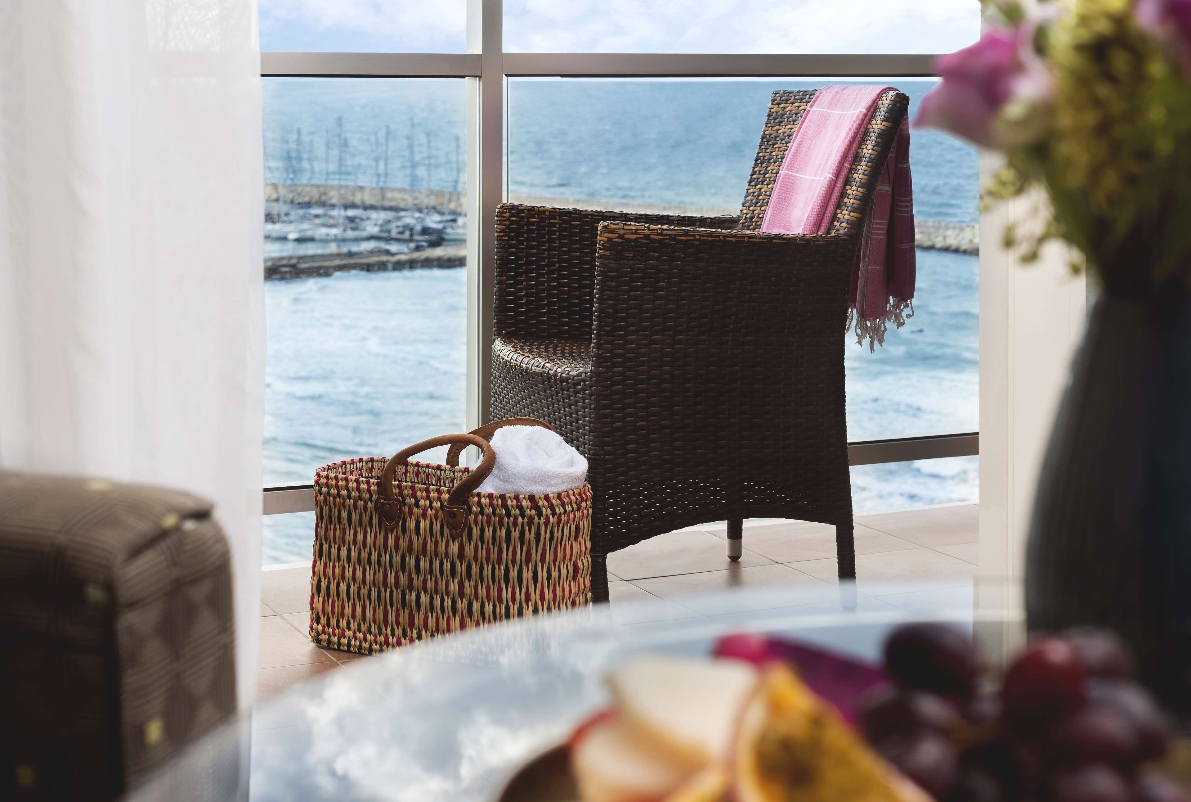 A wicker chair with a pink blanket, a woven bag, and a view of the sea through large windows.