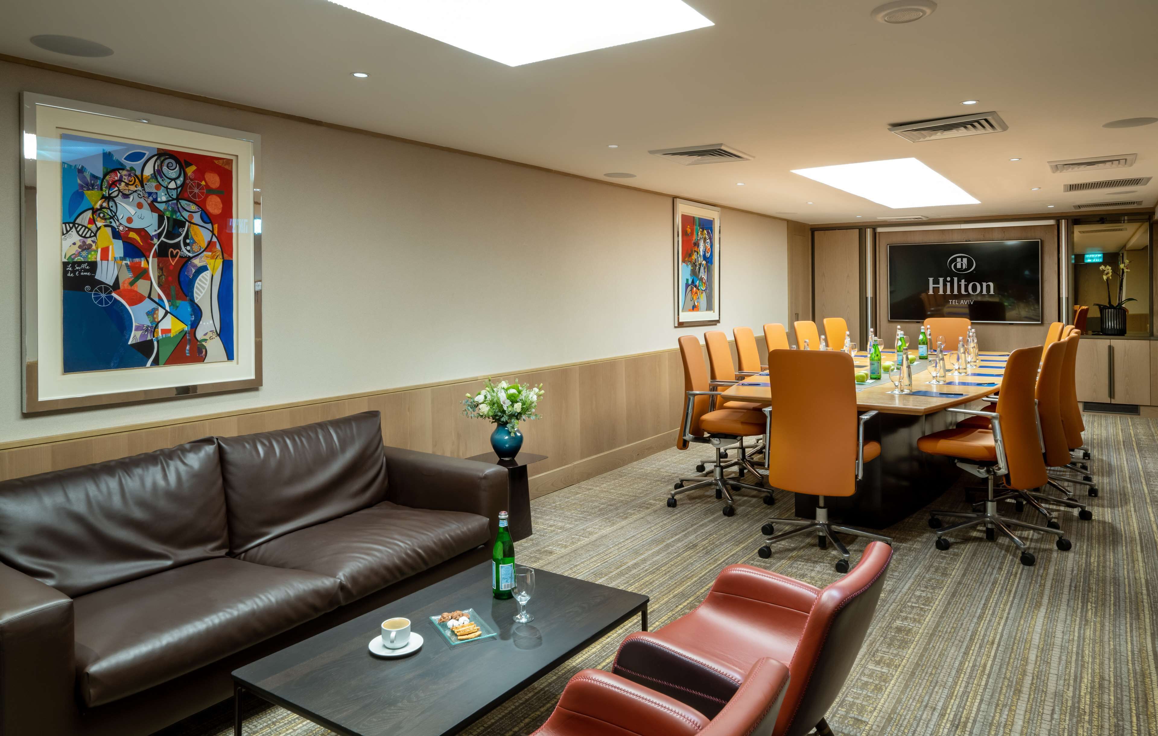 A conference room with a long table, orange chairs, a brown leather sofa, and a coffee table with a vase and a glass.