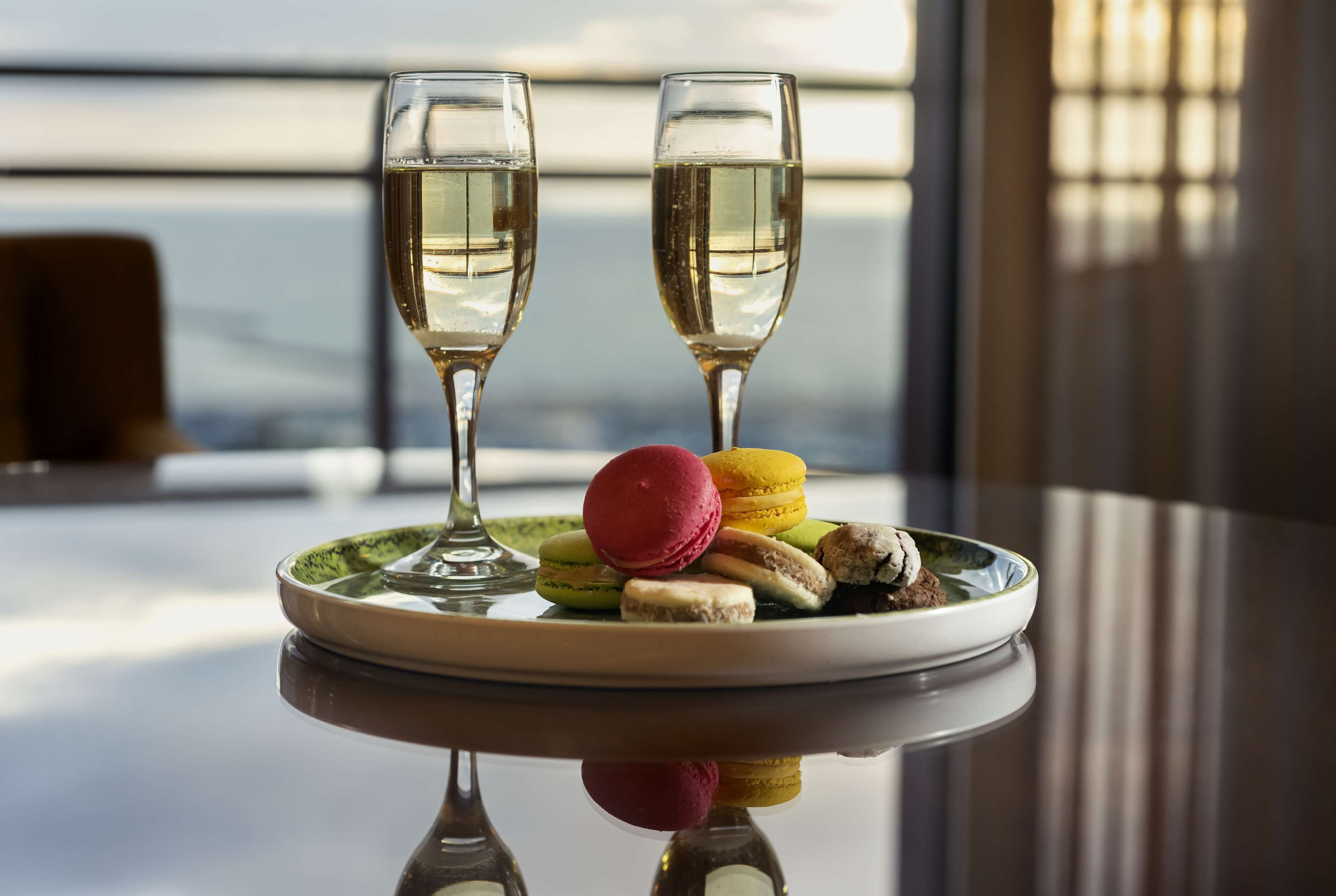 A table with two glasses of white wine, a plate of colorful macarons, and a dessert.