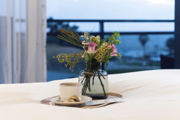 Una cama con sábana blanca, un florero con flores, una taza de café y una bandeja con un plato.