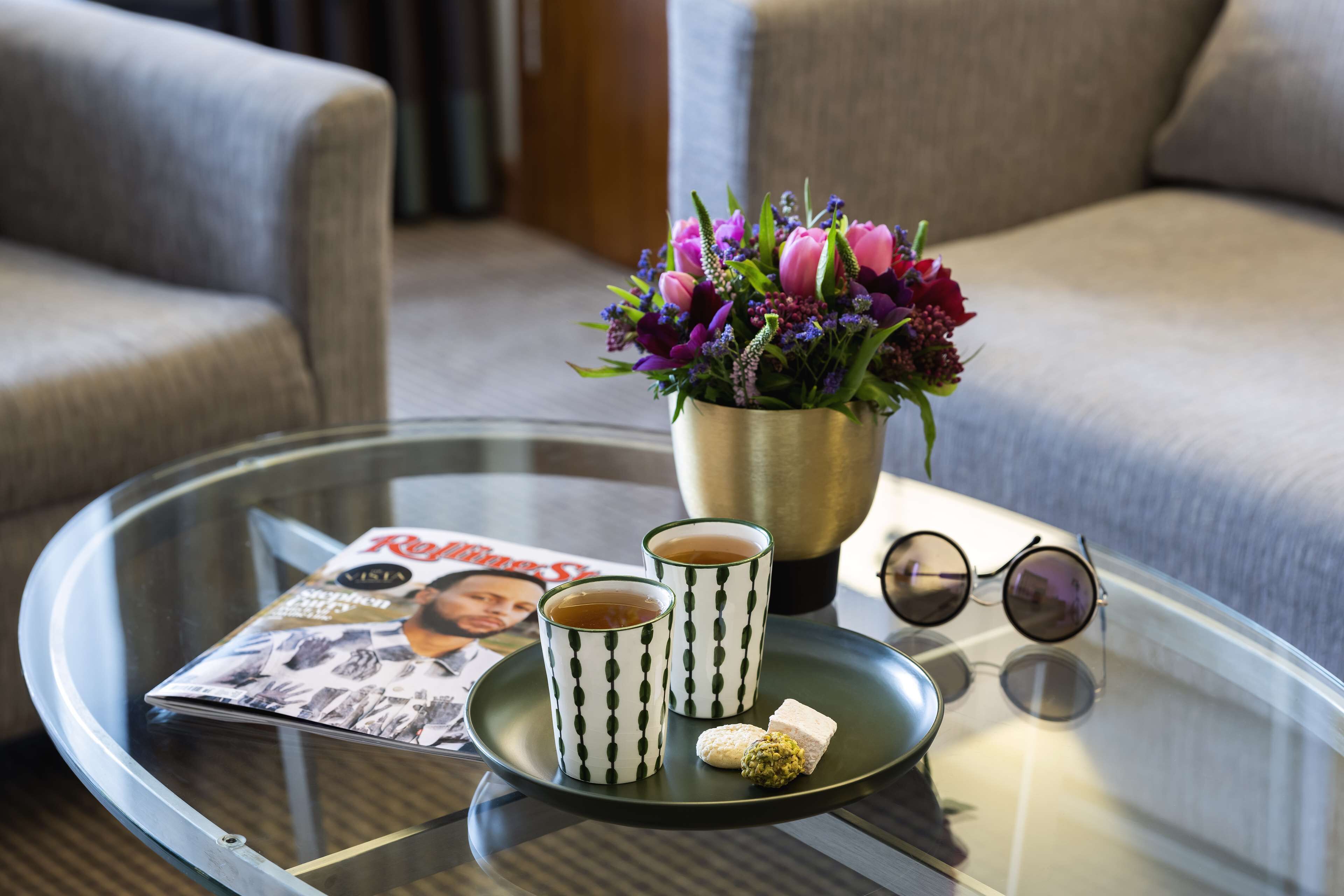 A glass table with a magazine, two cups of tea, a small plate with a pastry, a vase of flowers, and a pair of sunglasses.