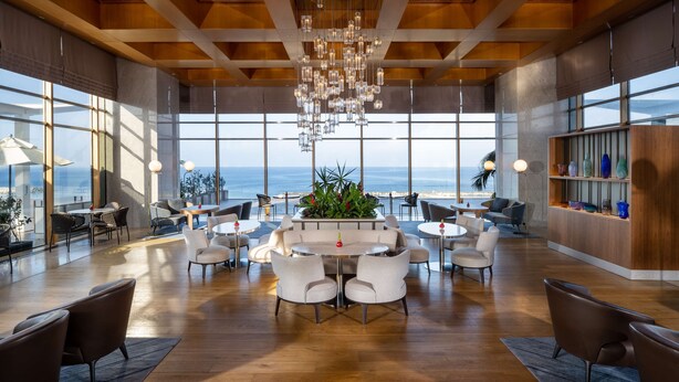 A spacious indoor area with a central seating arrangement, large windows offering a view of the ocean, and a wooden ceiling structure. - Hilton Tel Aviv (Tel Aviv)